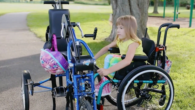 A girl in a wheelchair beside her blue Dream Bike in a park